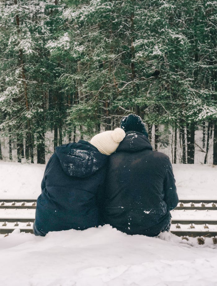 Couple in winter coats enjoy a snowy forest view sitting on railroad tracks.