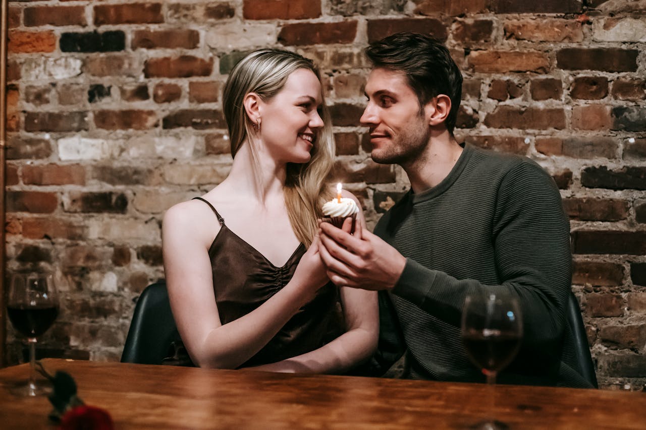 project-01-d Happy young couple in elegant clothes sitting at table and celebrating birthday with small cupcake and candle near glasses with red wine and brick wall while looking at each other
